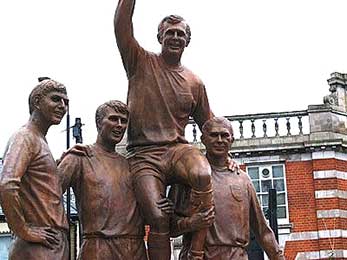 Op het beeld staan van links naar rechts Geoff Hurst, Martin Peters en Ray Wilson die aanvoerder Bobby Moore op de schouders dragen.